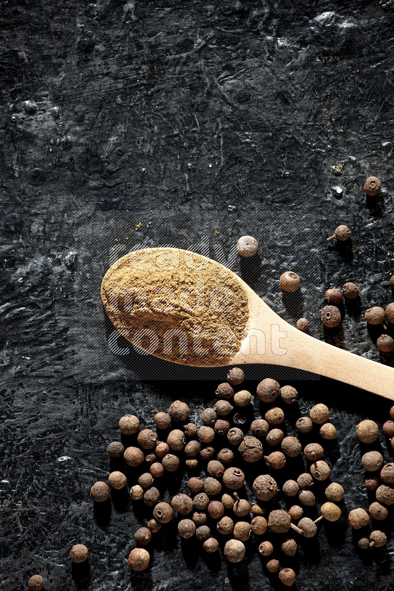 Wooden spoons full of all spice powder and allspice whole balls beside it on a textured black flooring
