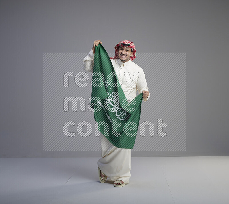 A saudi man standing wearing thob and red shomag holding big saudi flag on gray background