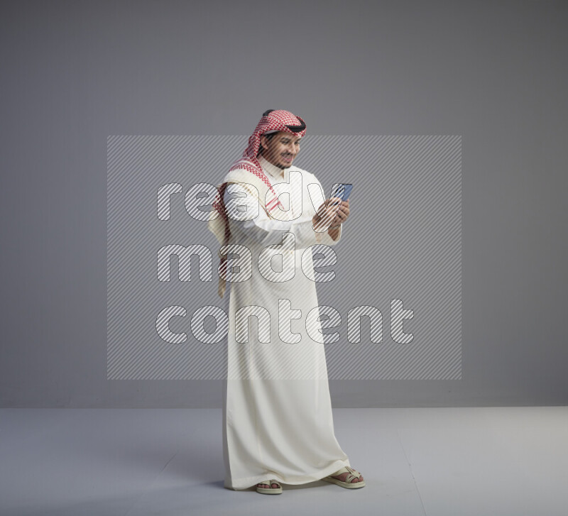 A  Saudi man standing wearing thob and red shomag texting on phone on gray background