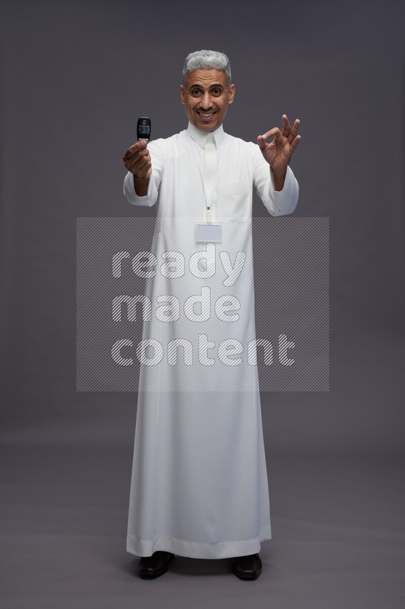Saudi man wearing thob with neck strap employee badge standing holding key car on gray background