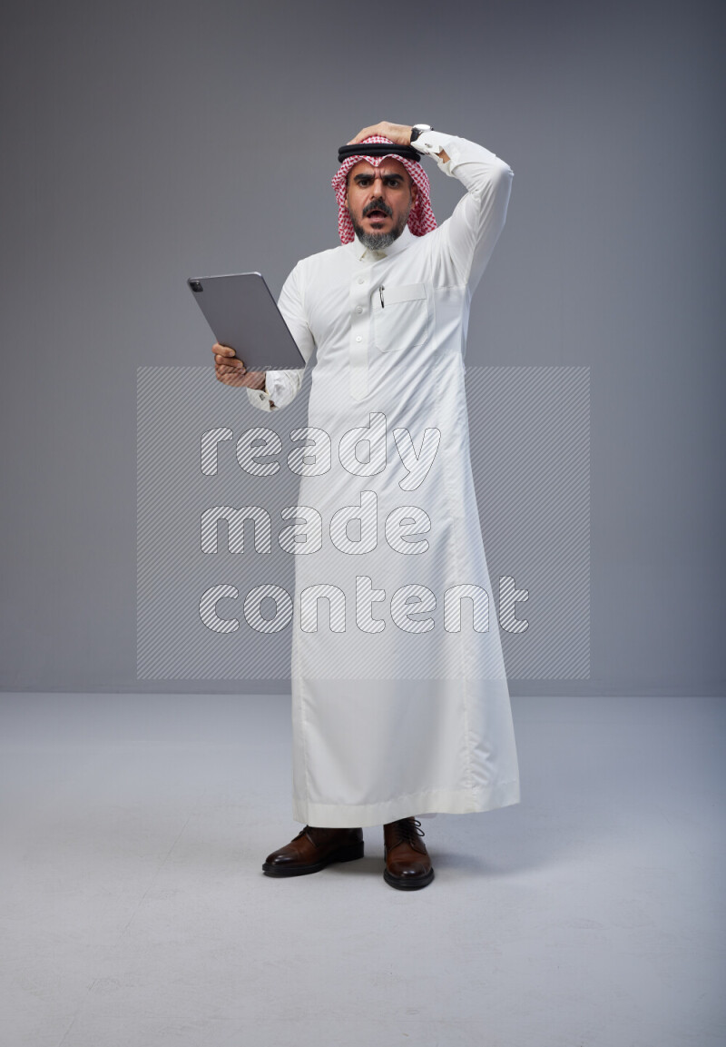 Saudi man Wearing Thob and red Shomag standing working on tablet on Gray background