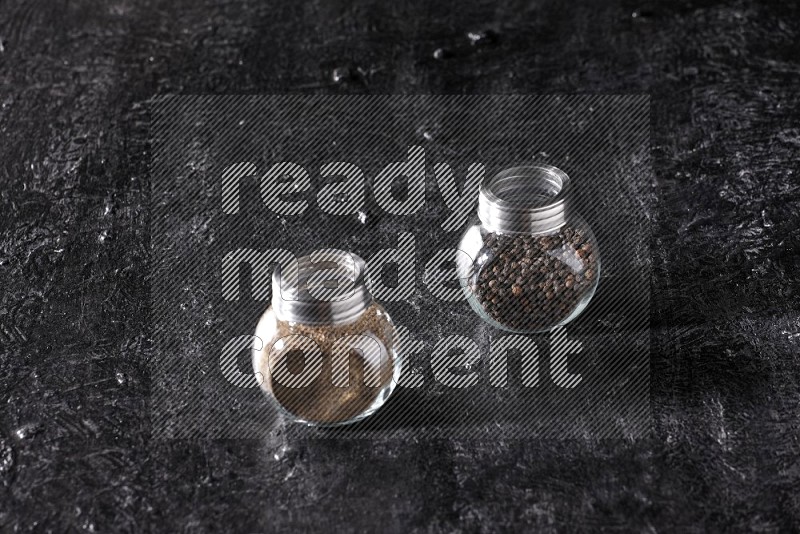 2 glass spice jars full of black pepper powder and black pepper beads on textured black flooring