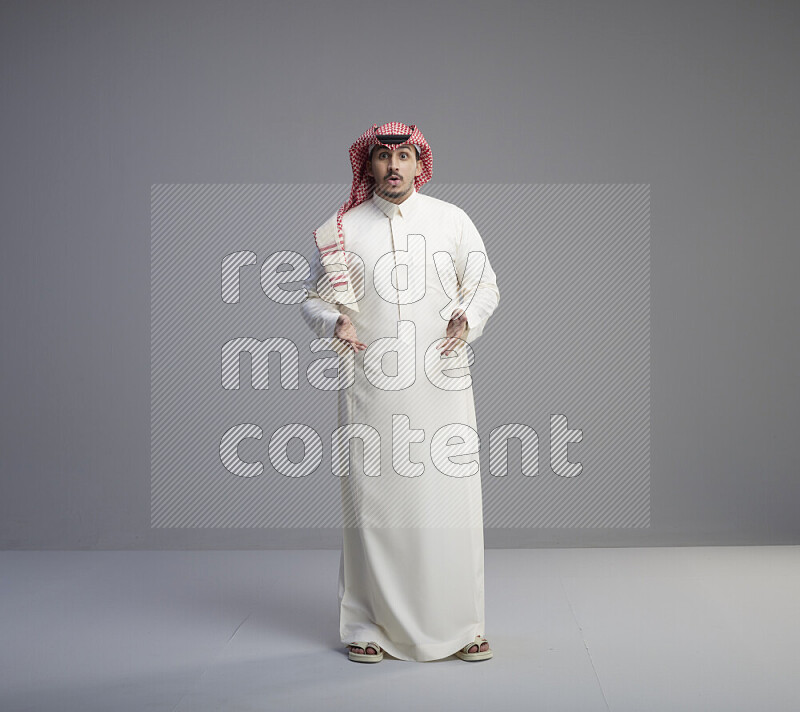 A Saudi man standing wearing thob and red shomag interacting with the camera on gray background