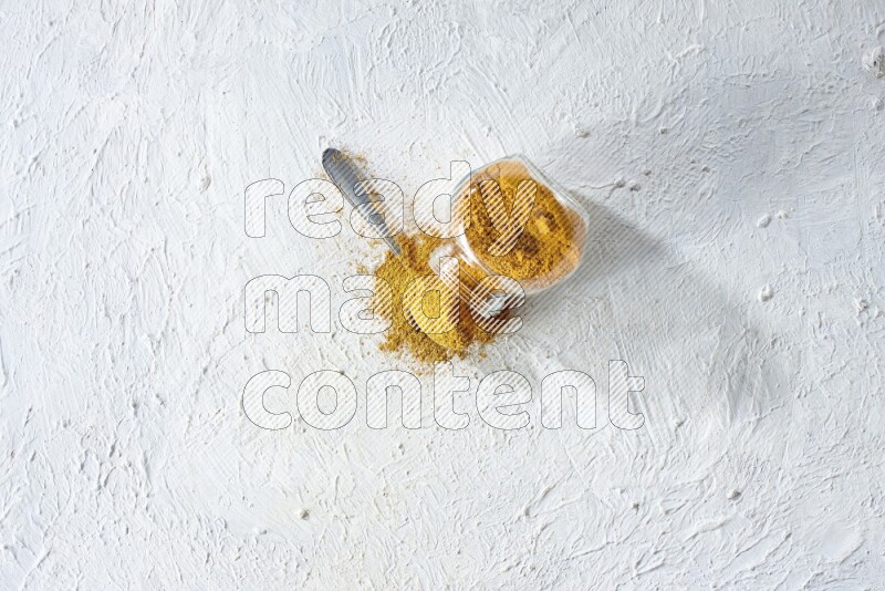 A flipped glass spice jar and a metal spoon full of turmeric powder and powder spilled out of it on textured white flooring