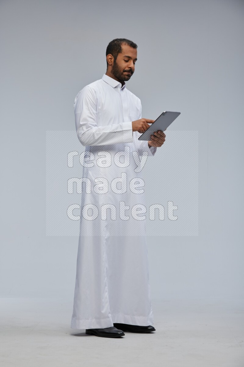Saudi man wearing Thob standing working on tablet on Gray background