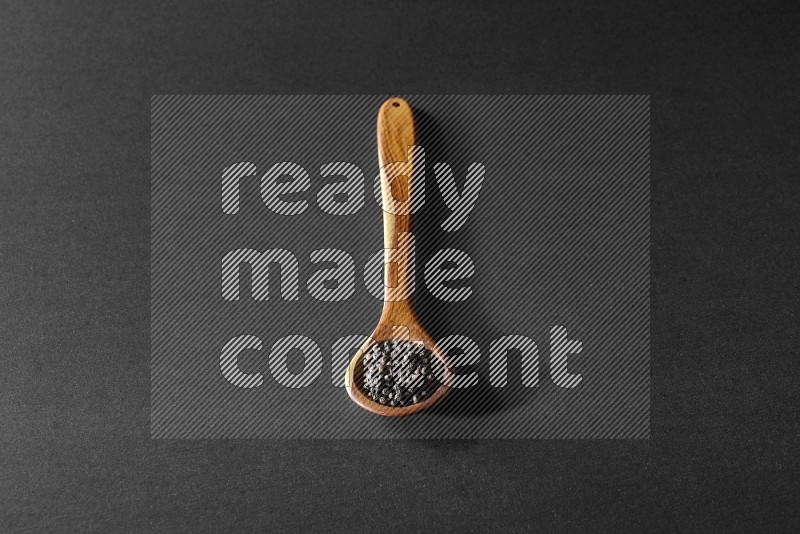 A wooden ladle full of black pepper beads on a black flooring