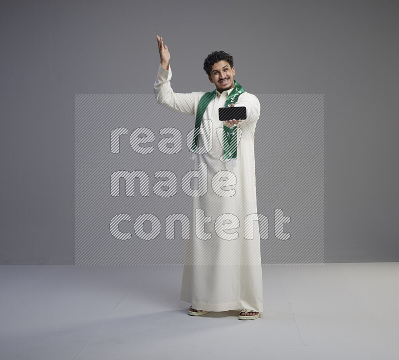 A Saudi man standing wearing thob and Saudi flag scarf showing phone to camera on gray background