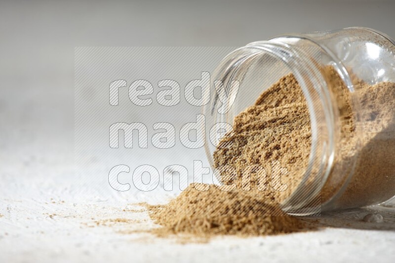 A flipped glass jar full of cumin powder and powder spilled out on textured white flooring
