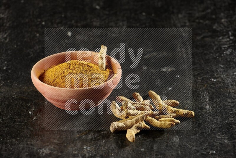 A wooden bowl full of turmeric powder with dried turmeric whole fingers on textured black flooring