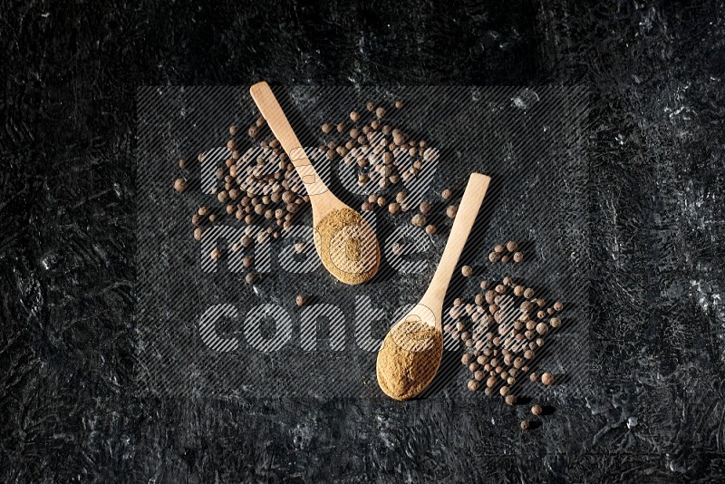 Wooden spoons full of all spice powder and allspice whole balls beside it on a textured black flooring