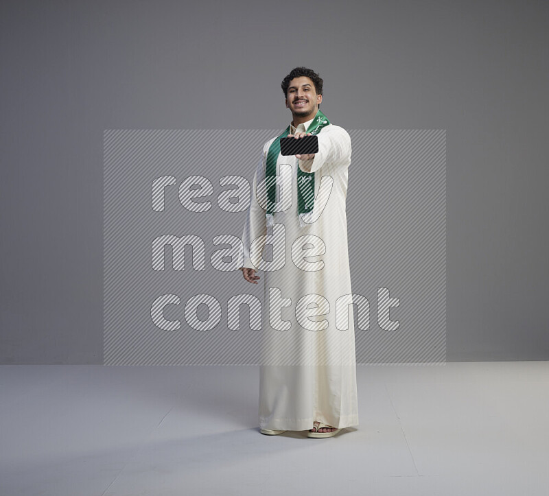 A Saudi man standing wearing thob and Saudi flag scarf showing phone to camera on gray background