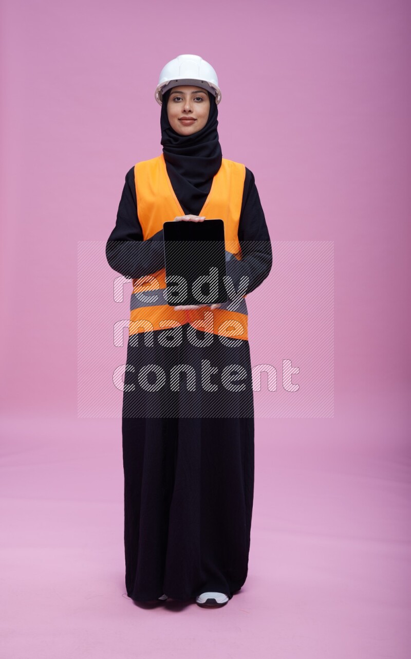 Saudi woman wearing Abaya with engineer vest and helmet standing showing tablet to camera on pink background