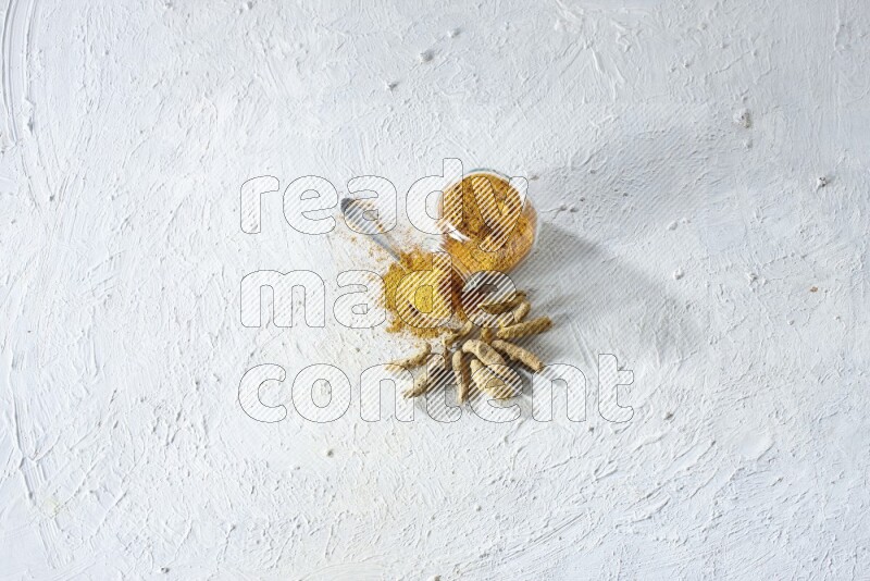 A flipped glass spice jar and metal spoon full of turmeric powder and powder spilled out of it with dried whole fingers on textured white flooring