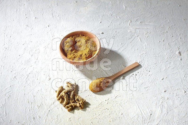A wooden bowl and wooden spoon full of turmeric powder with dried turmeric fingers on textured white flooring