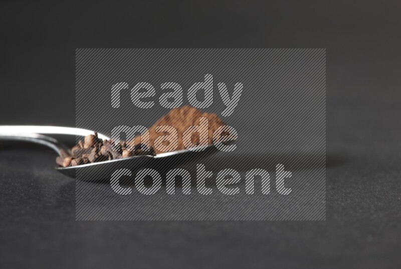 2 metal spoons, one full of cloves grains and the other full of cloves powder on a black flooring