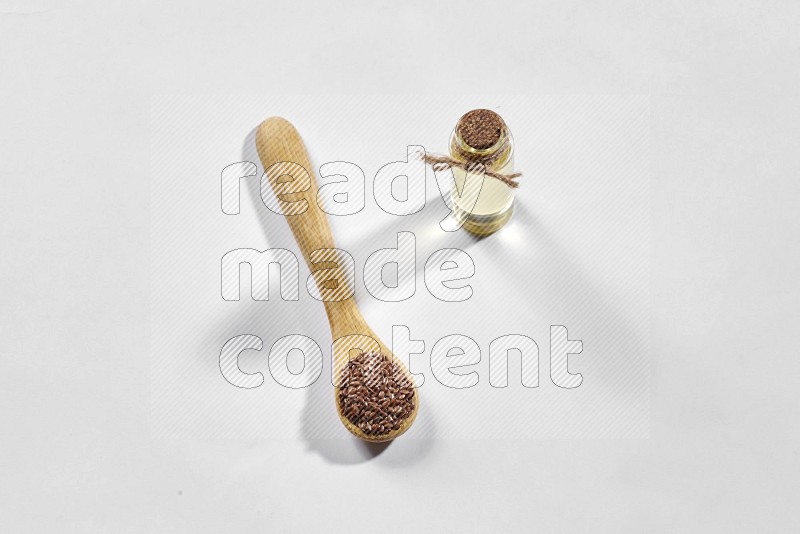 A wooden spoon full of flax seeds and a glass bottle full of flaxseeds oil on a white flooring