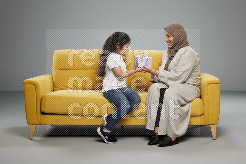 A girl sitting on a yellow sofa and giving a gift to her mother on gray background