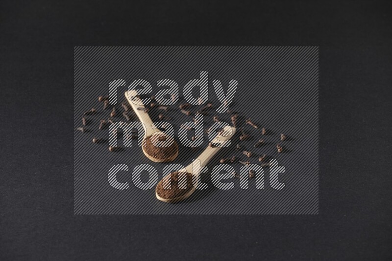 ملعقتان خشبيتان ممتلئتان ببودرة القرنفل مع حبوب القرنفل المتناثرة على خلفية سوداء