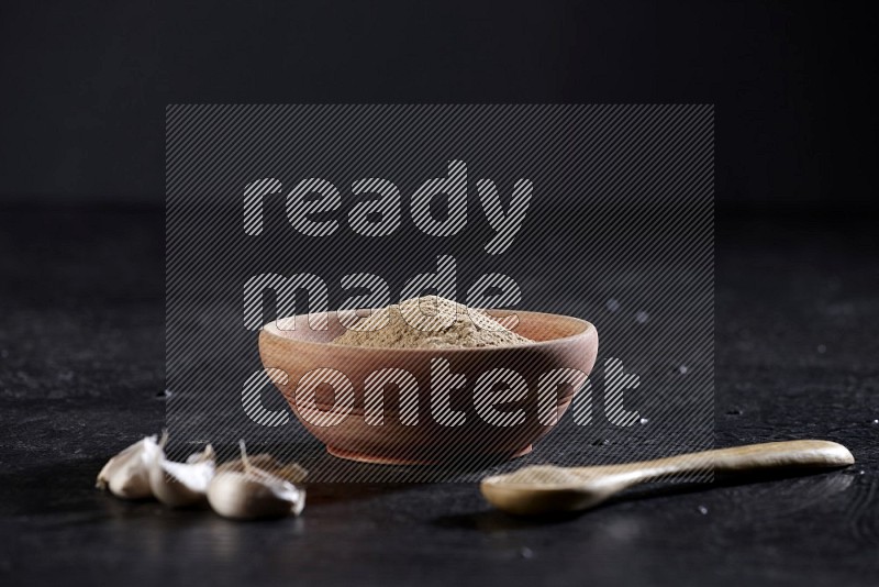 A wooden bowl and spoon full of garlic powder and beside it garlic cloves on a textured black flooring
