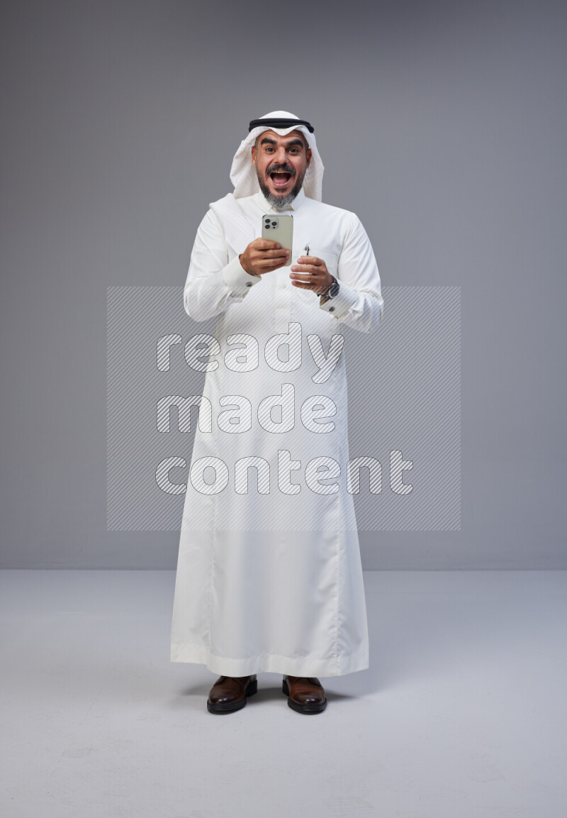 Saudi man Wearing Thob and white Shomag standing texting on phone on Gray background