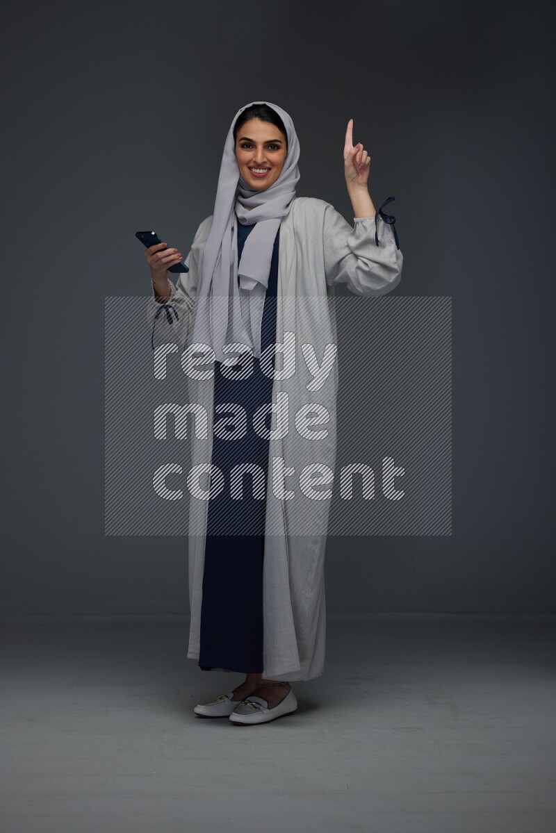 A Saudi woman wearing a light gray Abaya and head scarf standing and holding a phone on a grey background