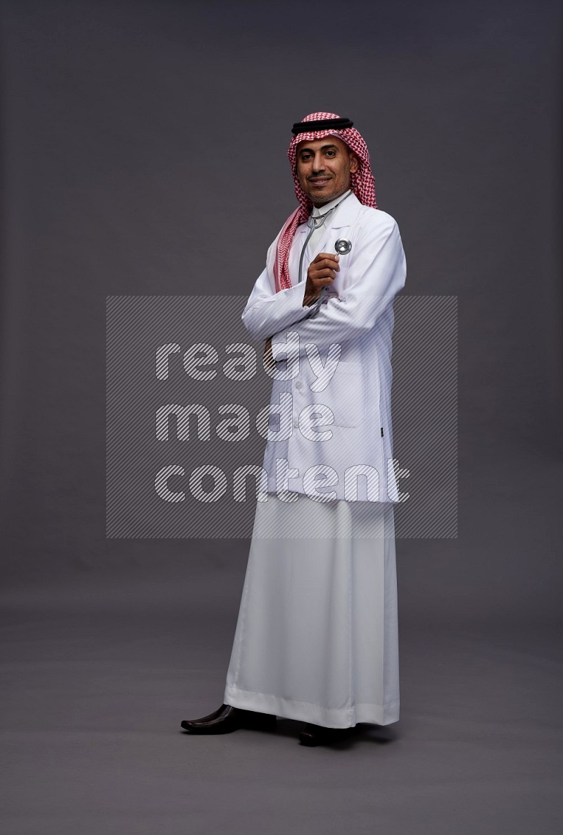 Saudi man wearing thob with lab coat and shomag with pocket employee badge with stethoscope standing interacting with the camera on gray background