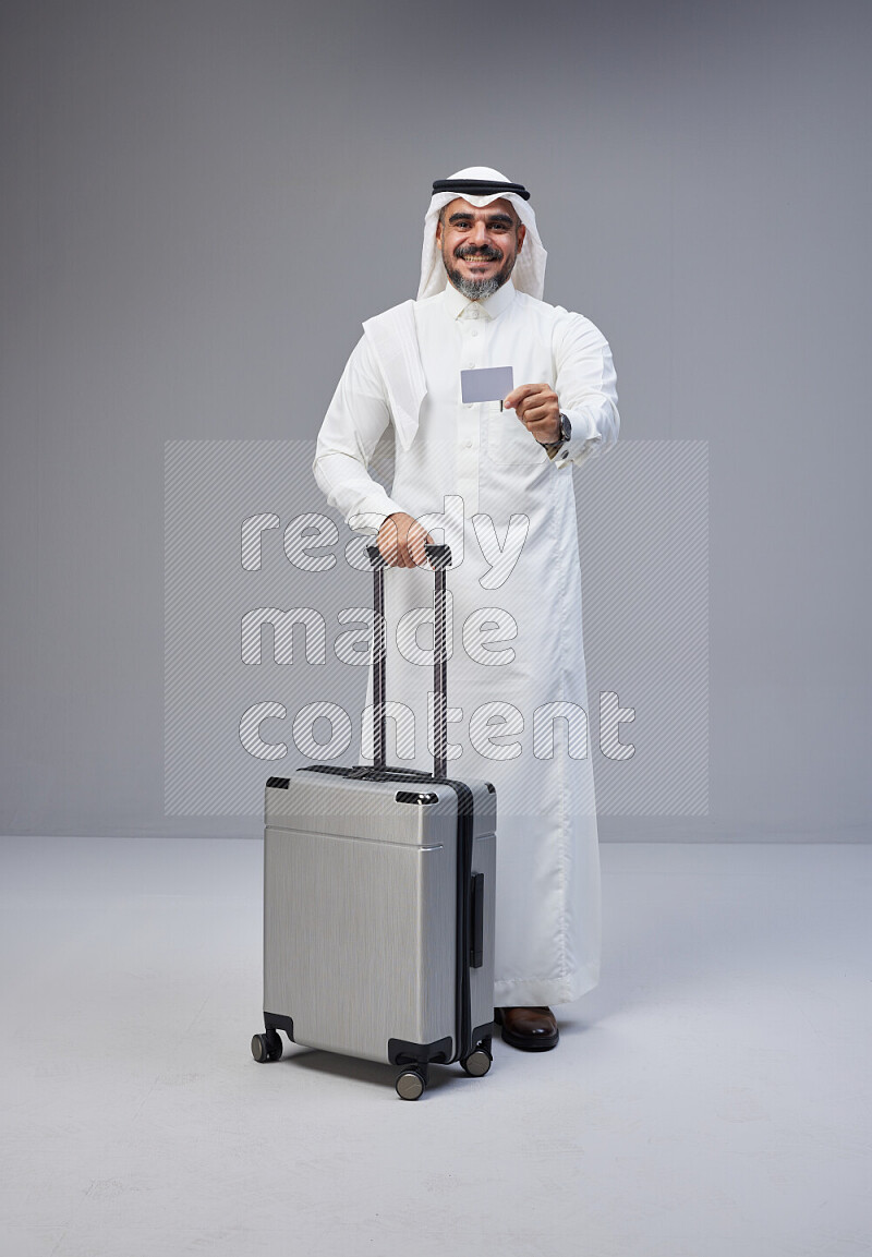 Saudi man wearing Thob and white Shomag standing holding Travel bag and ATM card on Gray background