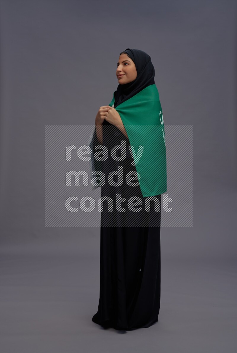 Saudi woman wearing Abaya standing holding Saudi flag on gray background
