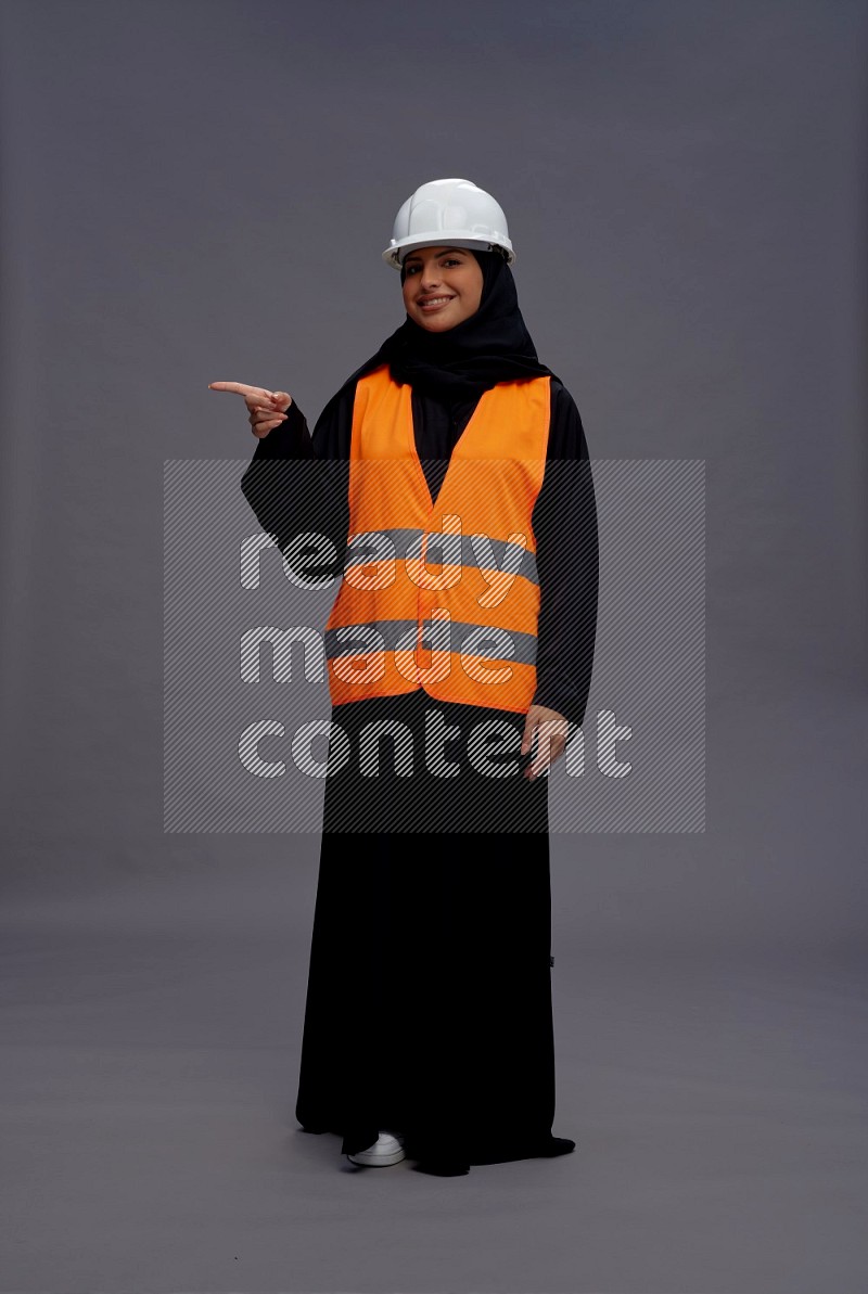 Saudi woman wearing Abaya with engineer vest standing interacting with the camera on gray background