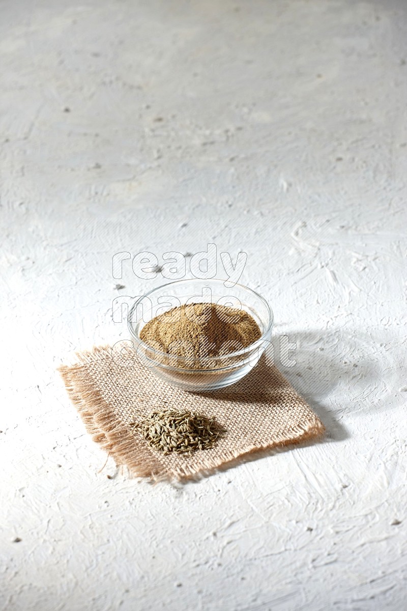 A glass bowl full of cumin powder with some of cumin seeds on burlap piece on a textured white flooring