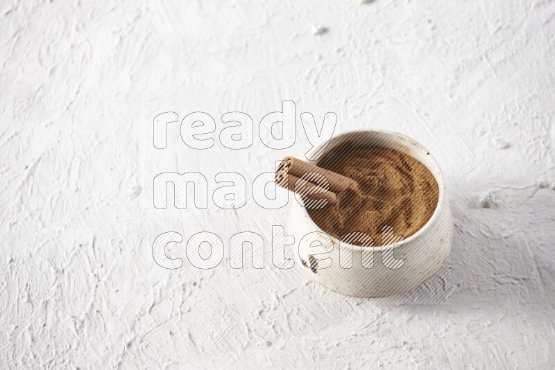 Ceramic beige bowl full of cinnamon powder with a cinnamon stick on a textured white background