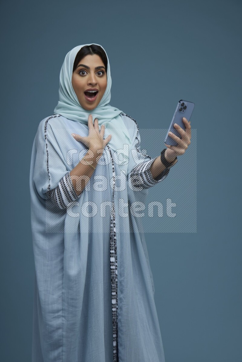 A Saudi woman posing with her phone in a blue background wearing a blue Abaya with hijab