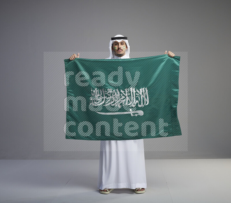 A Saudi man standing wearing thob and white shomag with face painting holding big Saudi flag on gray background