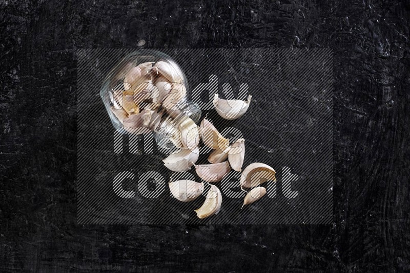 A glass spice jar full of garlic cloves flipped over with the cloves spilling out on a textured black flooring