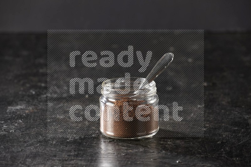 A glass jar full of cloves powder with a metal spoon on a textured black flooring