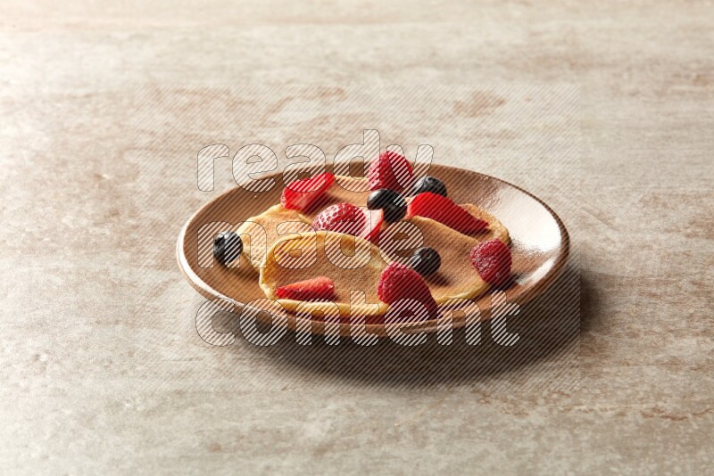 Five stacked mixed berries mini pancakes in a brown plate on beige background