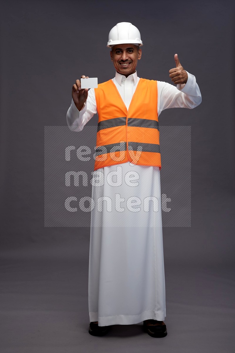 Saudi man wearing thob with engineer vest standing holding ATM card on gray background