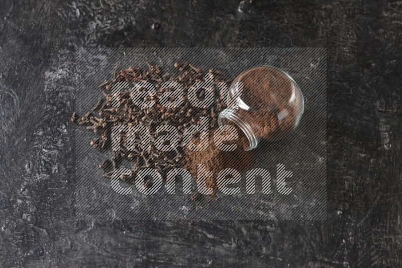 وعاء زجاجي للبهارات مقلوب ممتلئ ببودرة القرنفل والبودرة سقطت منه مع حبوب القرنفل المتناثرة على أرضية سوداء