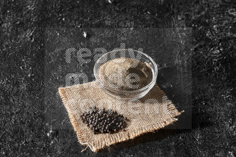 A glass bowl full of black pepper powder and black pepper beads on burlap fabric on textured black flooring