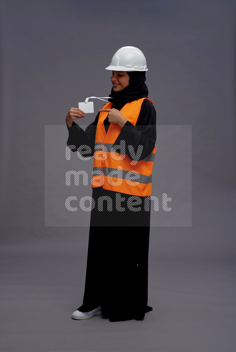 Saudi woman wearing Abaya with engineer vest with neck strap employee badge standing interacting with the camera on gray background