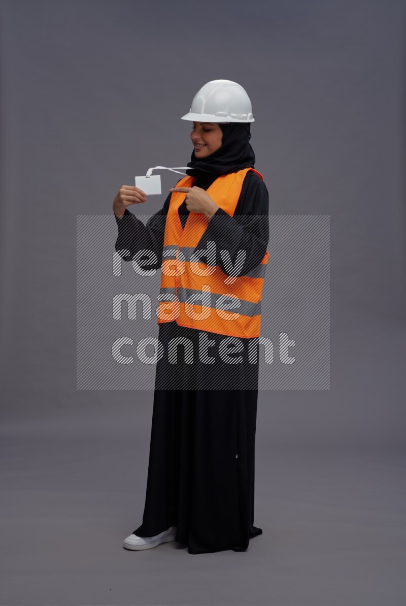 Saudi woman wearing Abaya with engineer vest with neck strap employee badge standing interacting with the camera on gray background