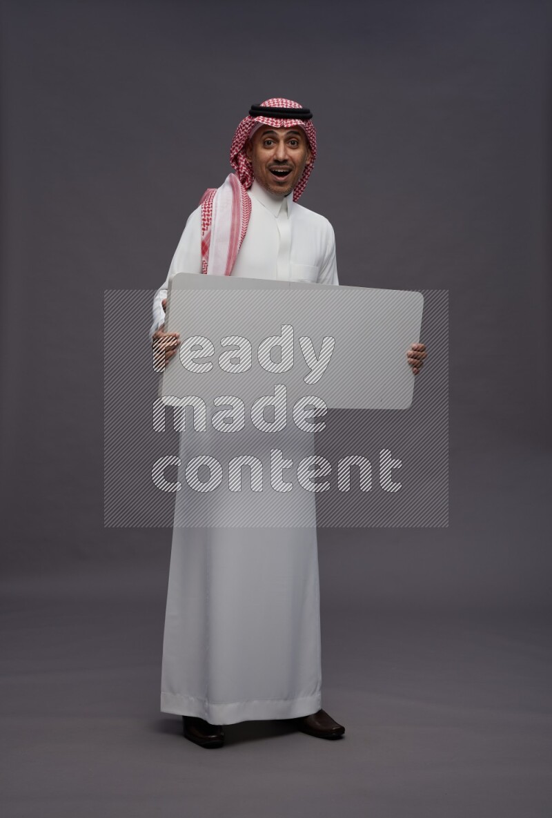 Saudi man wearing thob and shomag standing holding board on gray background