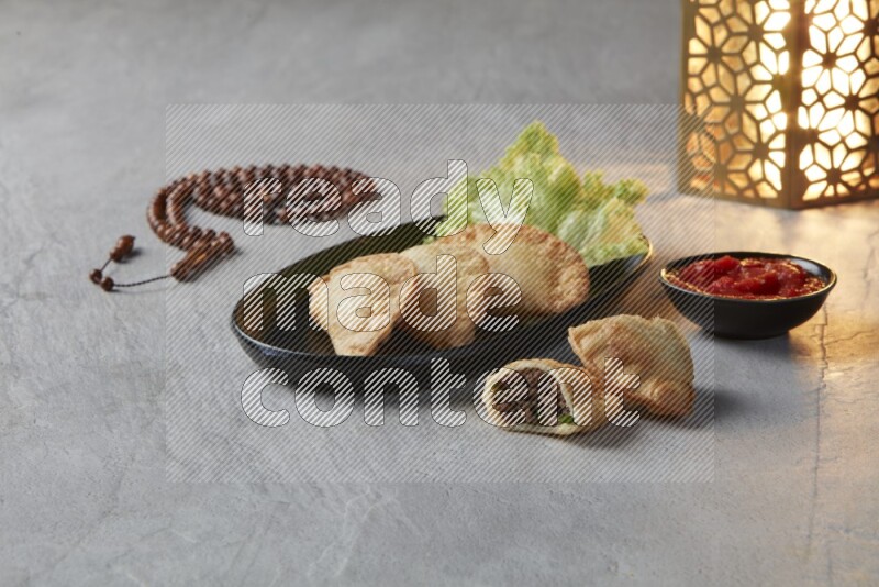 Three fried sambosas in an oval shaped black plate and a red sauce in a black round ramekin with a brown misbaha and a golden lantern on the side on a gray background