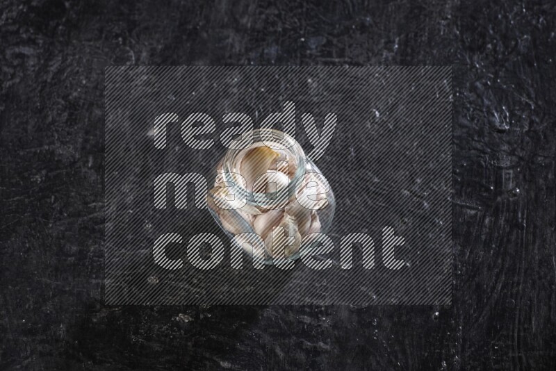 A glass spice jar full of garlic cloves on a textured black flooring
