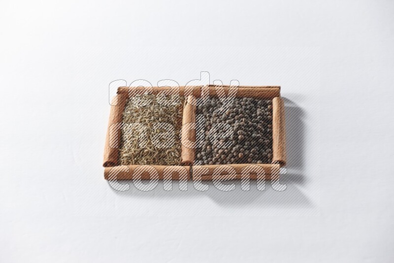 مربعان من أعواد القرفة ممتلئان بحبوب الفلفل الأسود وحبوب الكمون علي خلفية بيضاء