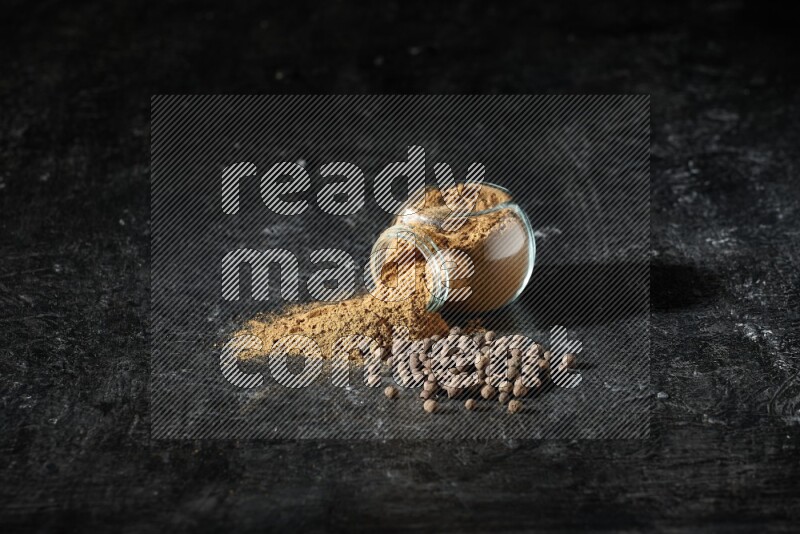 وعاء زجاجي للتوابل مقلوب ممتلئ ببودرة بهار الفلفل الحلو والبودرة تناثرت منه مع الحبوب على خلفية سوداء