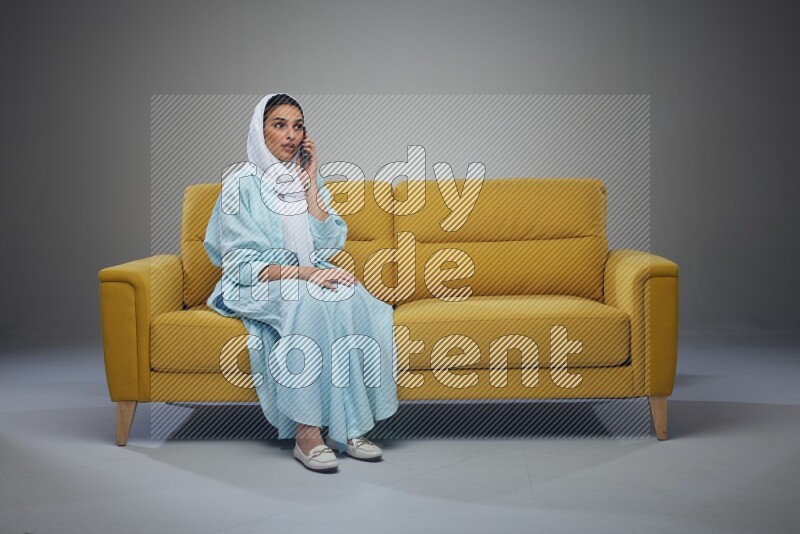 A Saudi woman wearing a light blue Abaya and a white head scarf sitting on a yellow sofa and talking in the phone while making different poses eye level on a grey background