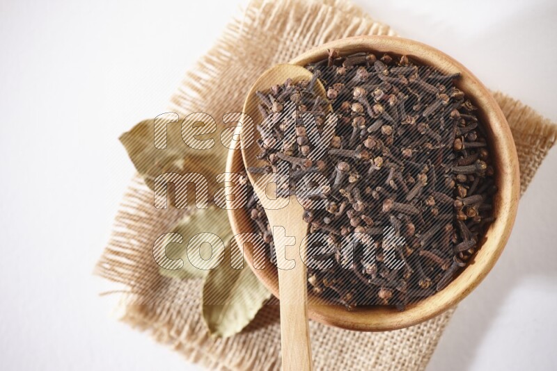 A wooden bowl and a wooden spoon full of cloves on a piece of burlap on a white flooring