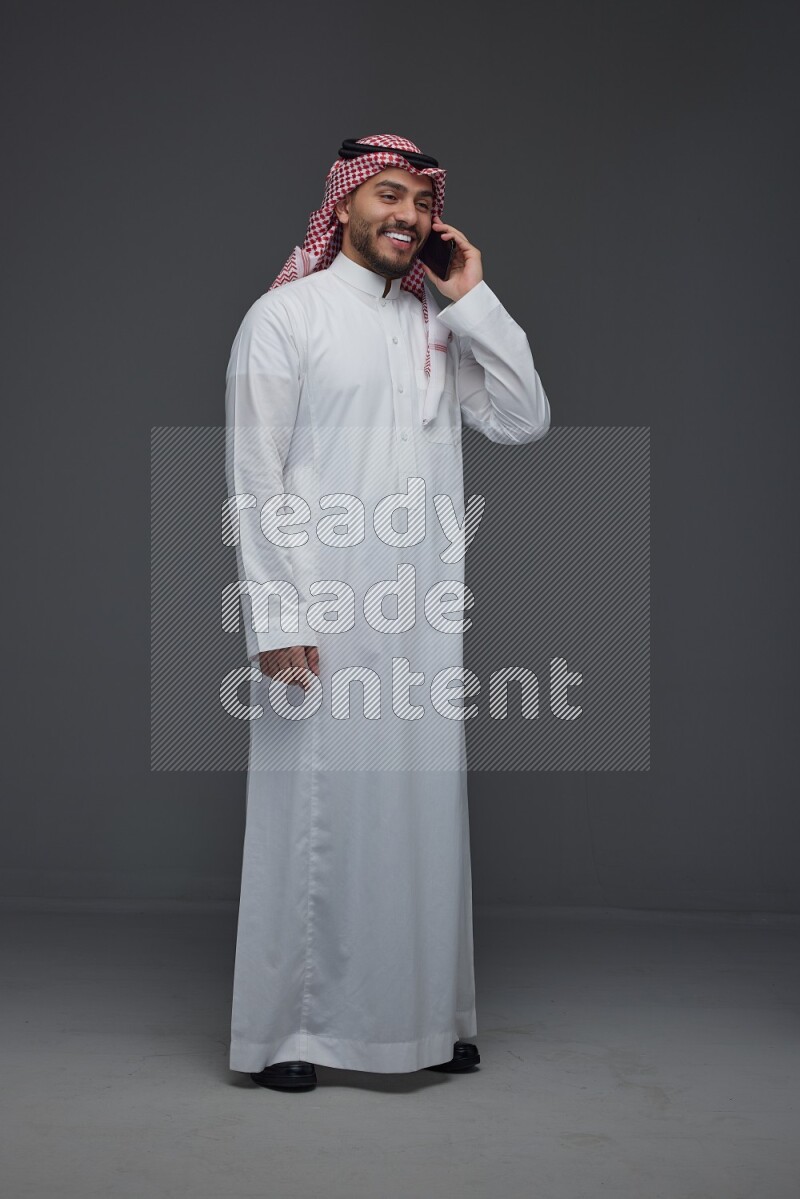 A Saudi man wearing Thobe and Shmagh talking in the phone eye level on a gray background