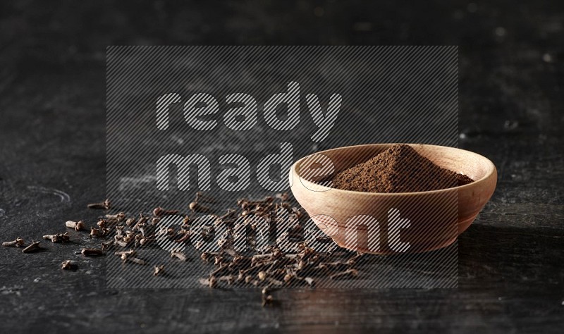 A wooden bowl full of cloves powder on a textured black flooring